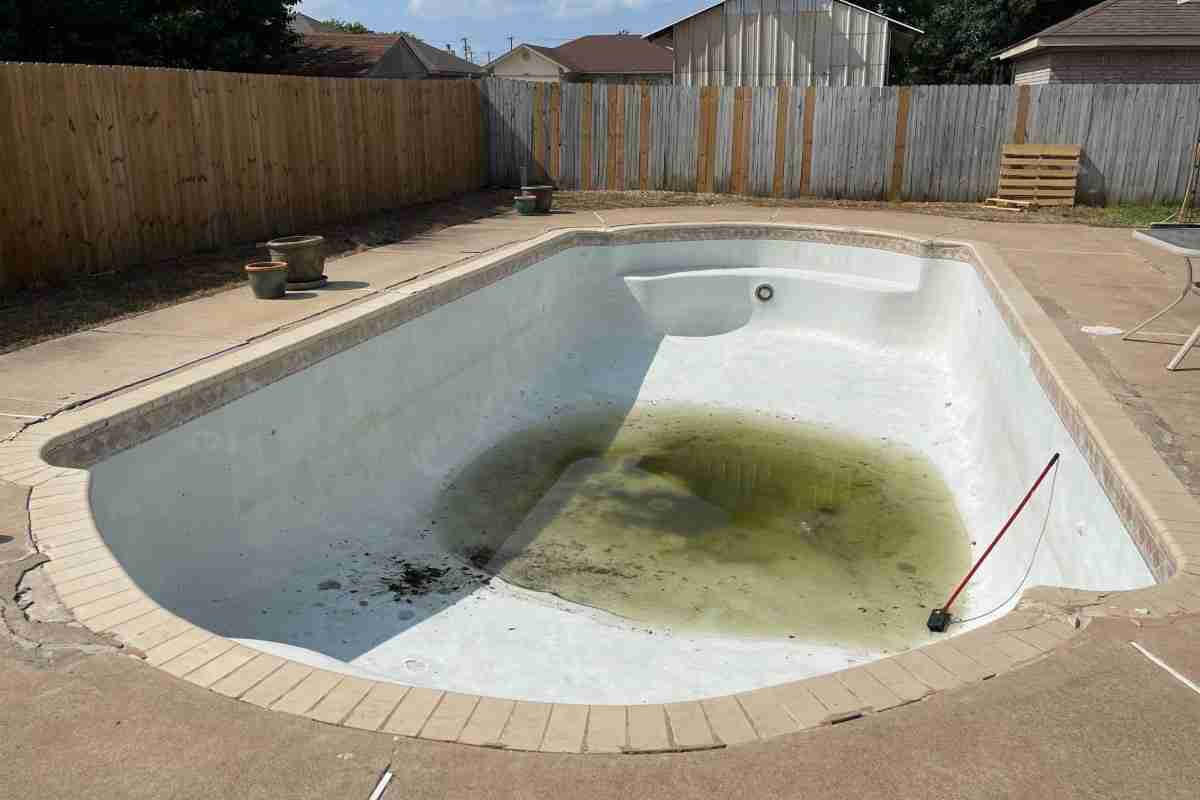 An empty swimming pool with a large, stagnant puddle of greenish water at the bottom, surrounded by a wooden fence and sparse outdoor furniture—a stark reminder to call trusted pool contractors for maintenance.