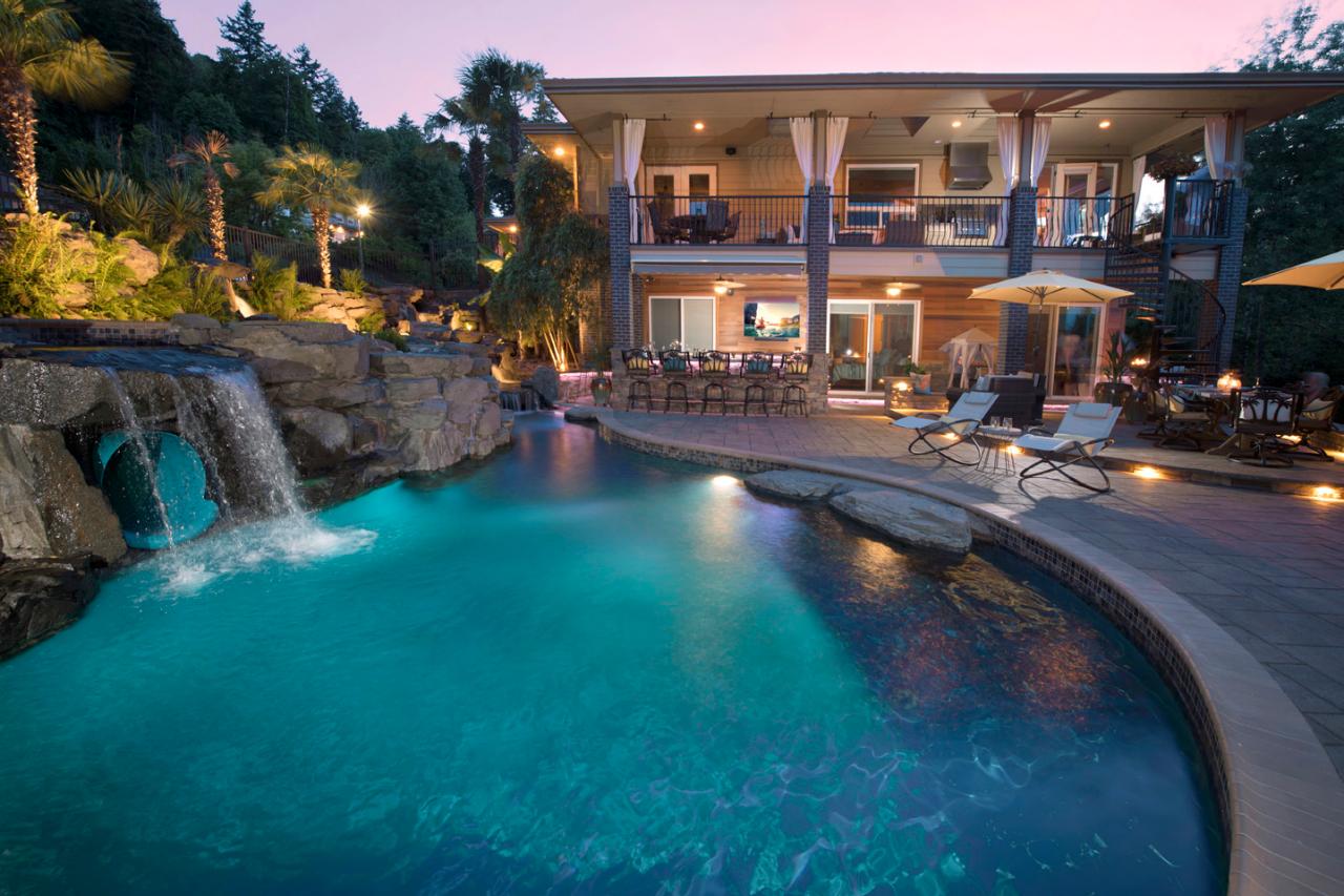 A two-story house with a balcony, spa-like pool, and waterfall feature in the backyard at dusk. The pool area has lounge chairs and outdoor seating with ambient lighting. Trees and greenery surround the property, creating a serene patio oasis.