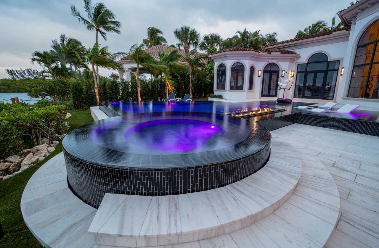 Luxury modern house with an infinity pool, illuminated purple jacuzzi, lounge chairs, and palm trees in the background. The setting is outdoors with a clear view of the sky and greenery.
